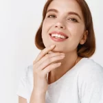 Smiling young woman with hand near chin, wearing white top.