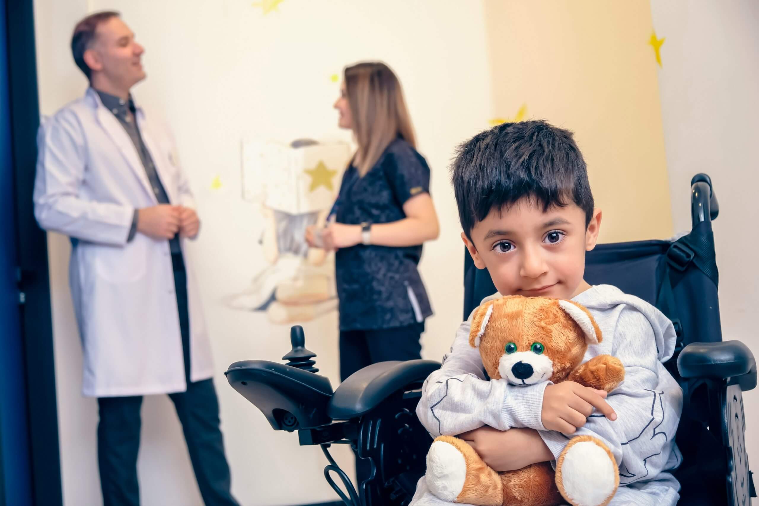 A young boy in a wheelchair holding a teddy bear, with adults talking in the background.