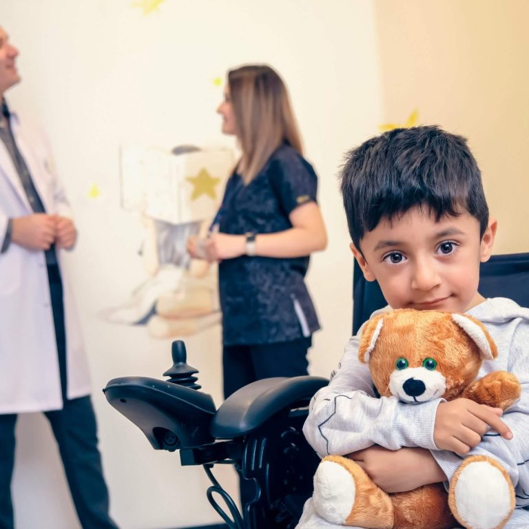 A young boy in a wheelchair holding a teddy bear, with adults talking in the background.
